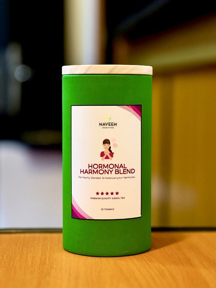 Green cylindrical container of Naveen House of Teas Hormonal Harmony Blend placed on a wooden table, with a soft-focus indoor background highlighting its natural, premium packaging.