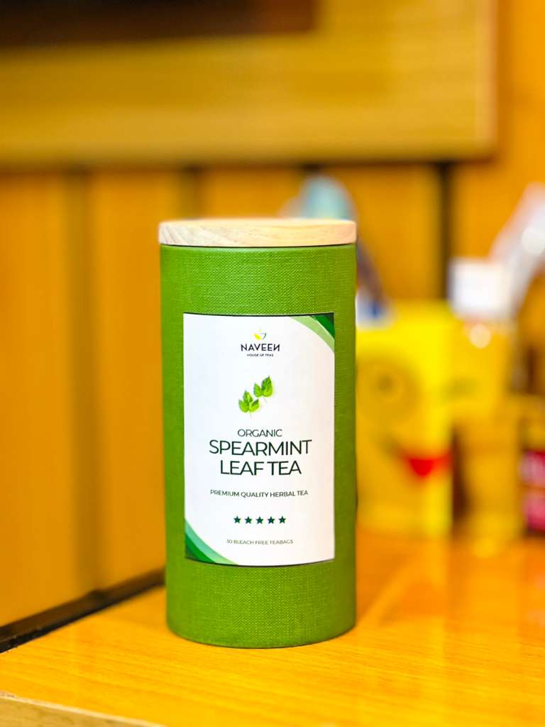 A green cylindrical container of Naveen Organic Spearmint Leaf Tea placed on a polished wooden table with a softly blurred yellow background.
